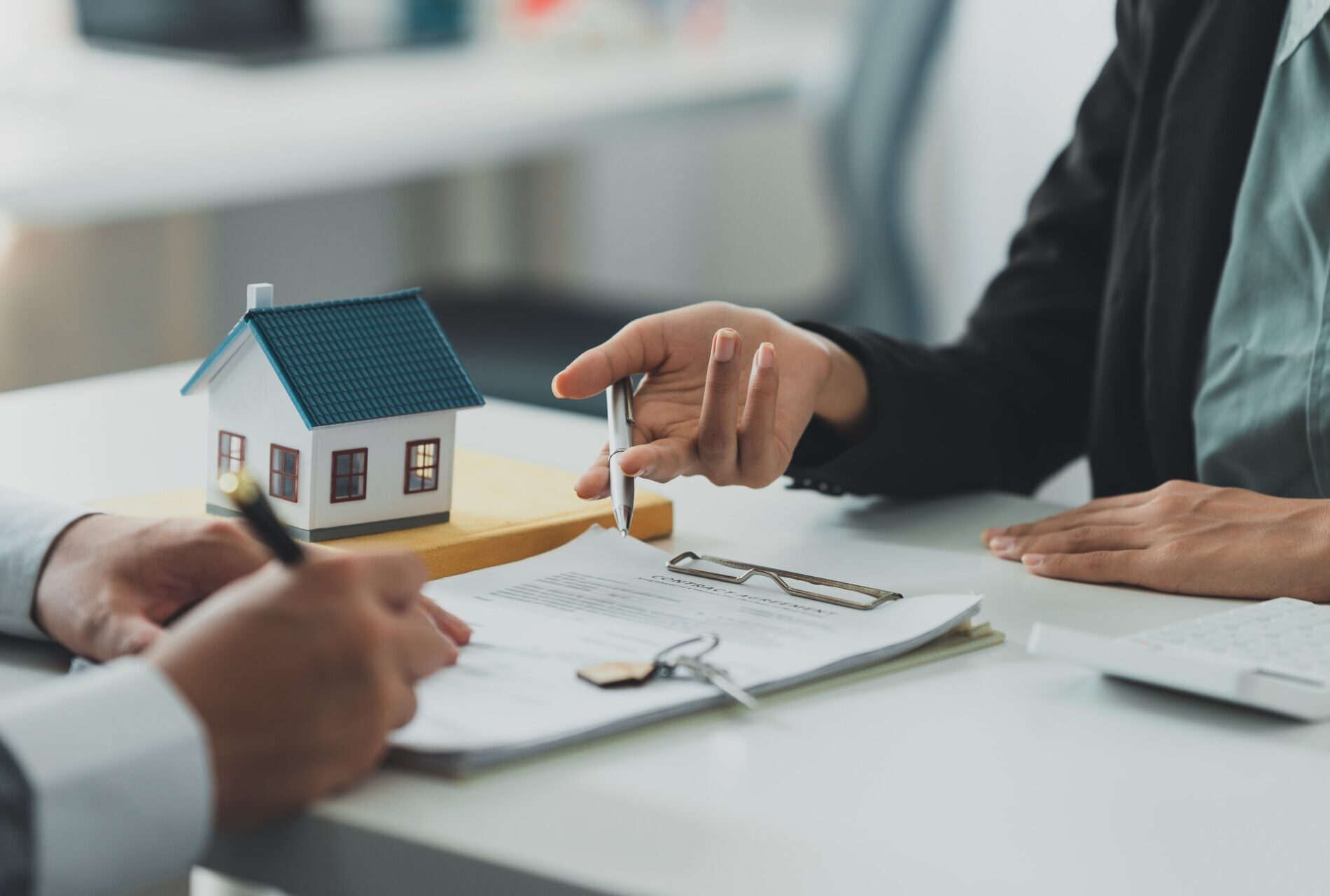 Ein Immobilienmakler zeigt mit einem Stift auf einen Vertrag, während ein Kunde kurz davor ist, ihn zu unterzeichnen. In der Mitte des Tisches stehen ein kleines Modellhaus und ein Schlüsselbund auf den Dokumenten.